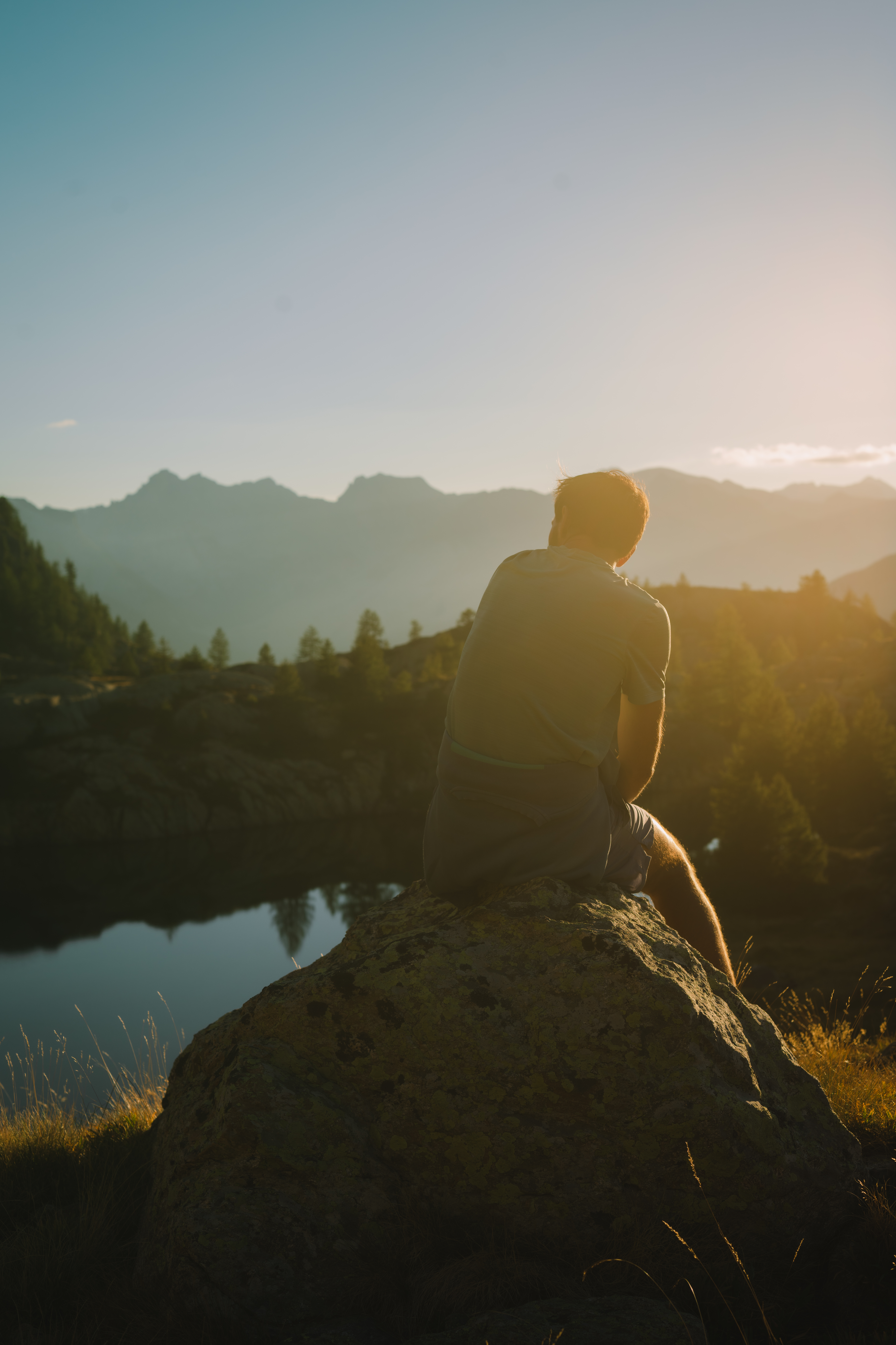 Un homme assis sur un rocher, regardant un paysage de montagnes au coucher du soleil, près d'un lac.