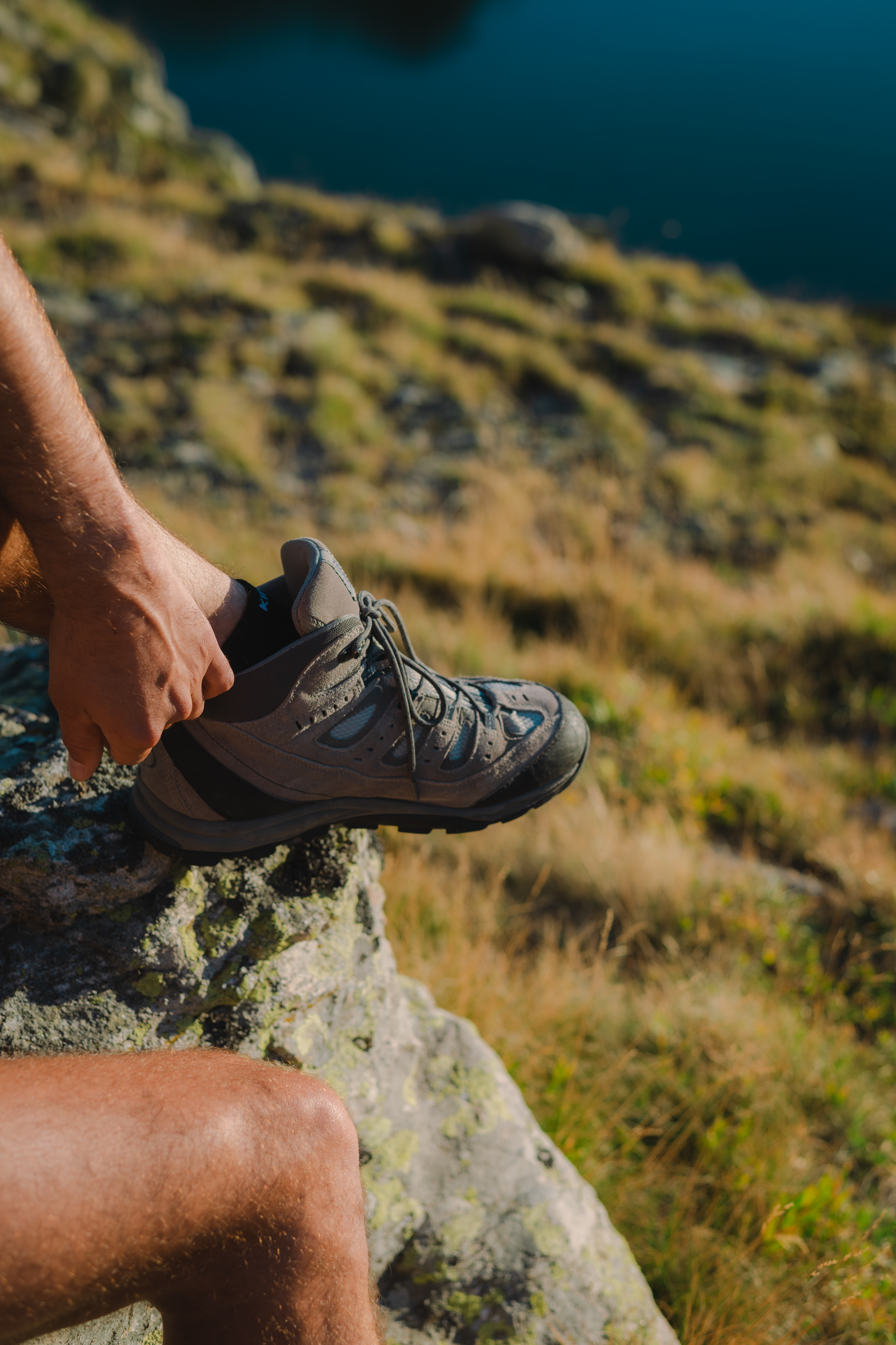 Un homme en train de lacer une chaussure de randonnée sur une pierre, avec un paysage de montagne en arrière-plan.