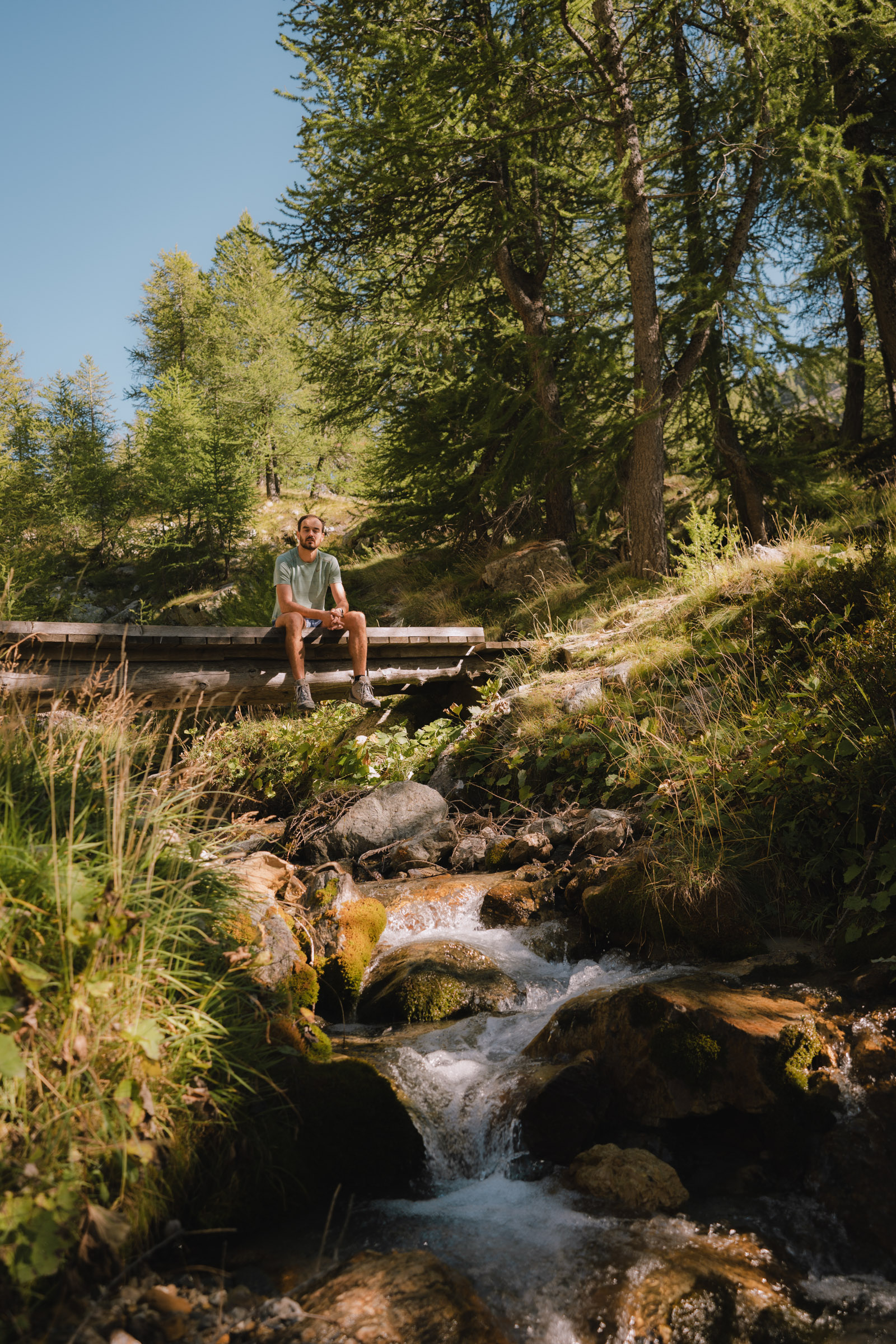 Un homme assis sur un pont en bois au-dessus d'un ruisseau, entouré d'arbres verts et d'une nature luxuriante.
