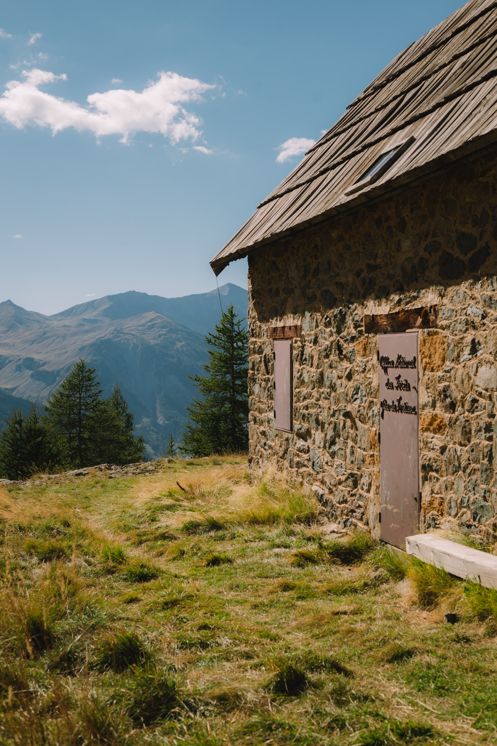 Un chalet en pierre dans les montagnes, avec un ciel bleu parsemé de nuages et des collines verdoyantes en arrière-plan.