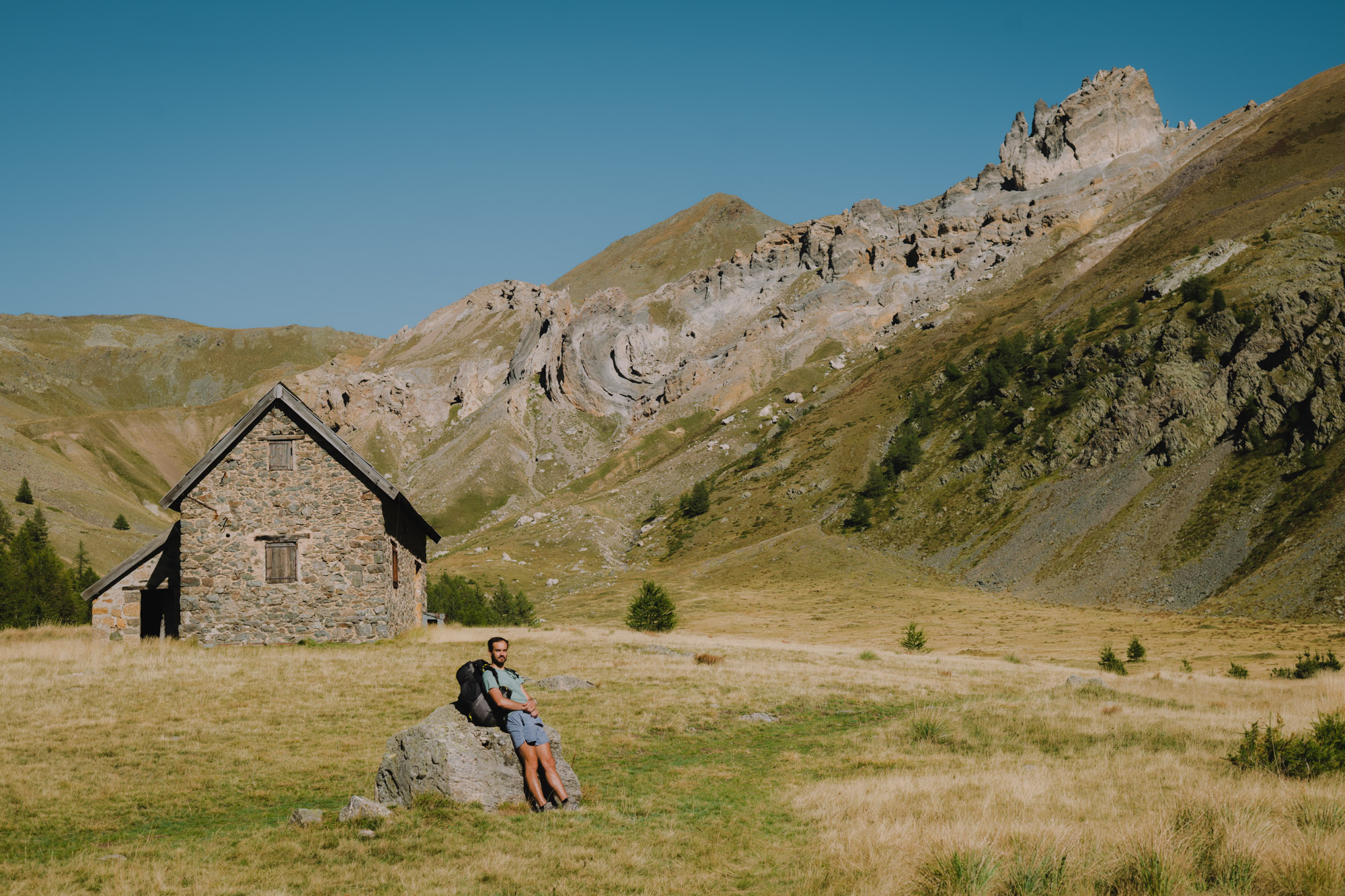 Un homme assis sur une pierre à côté d'une maison en pierre, avec des montagnes en arrière-plan et un ciel bleu dégagé.