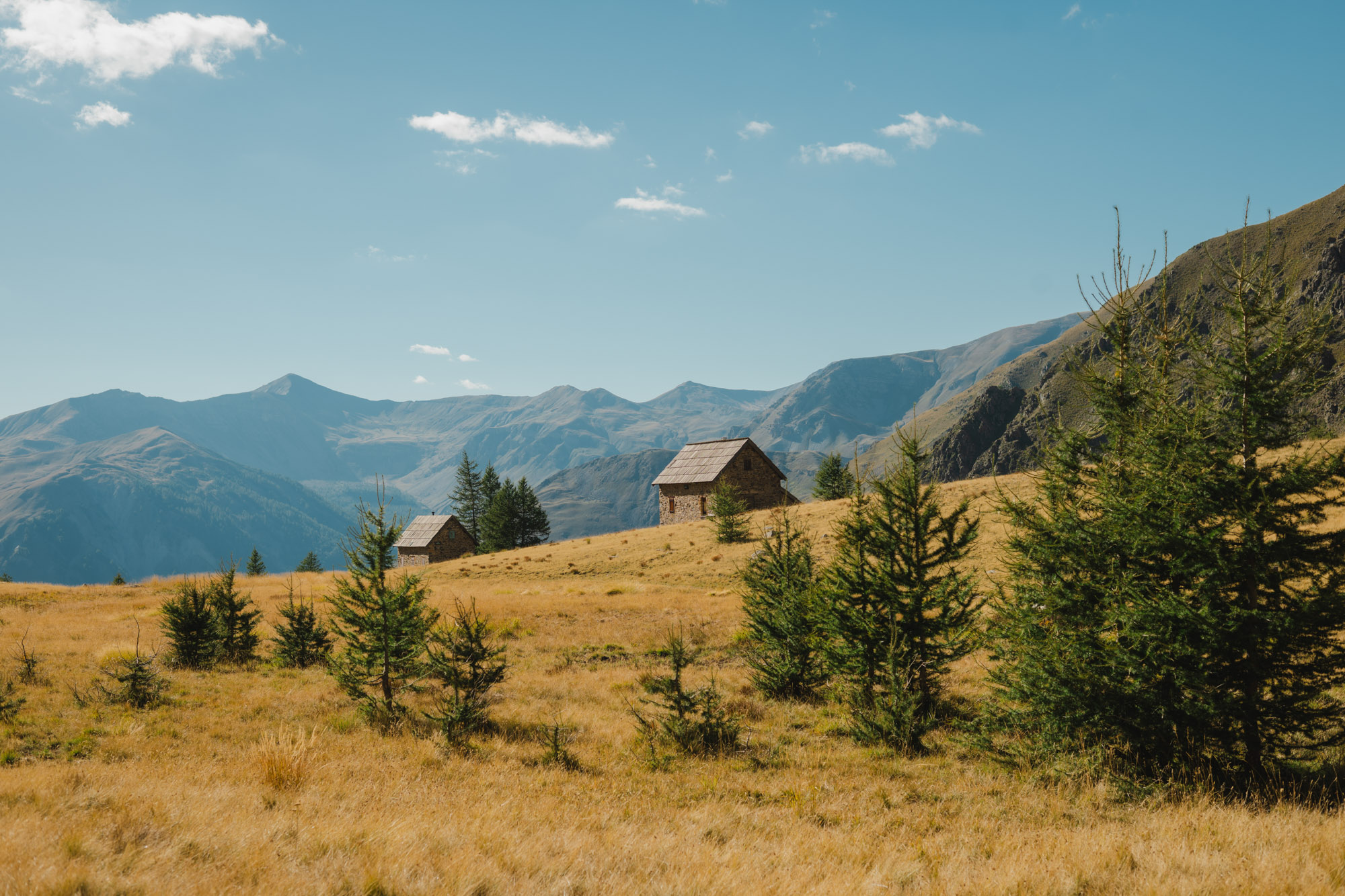 Deux petites cabanes en pierre sur une prairie dorée, entourées d'arbres et de montagnes au loin sous un ciel bleu.