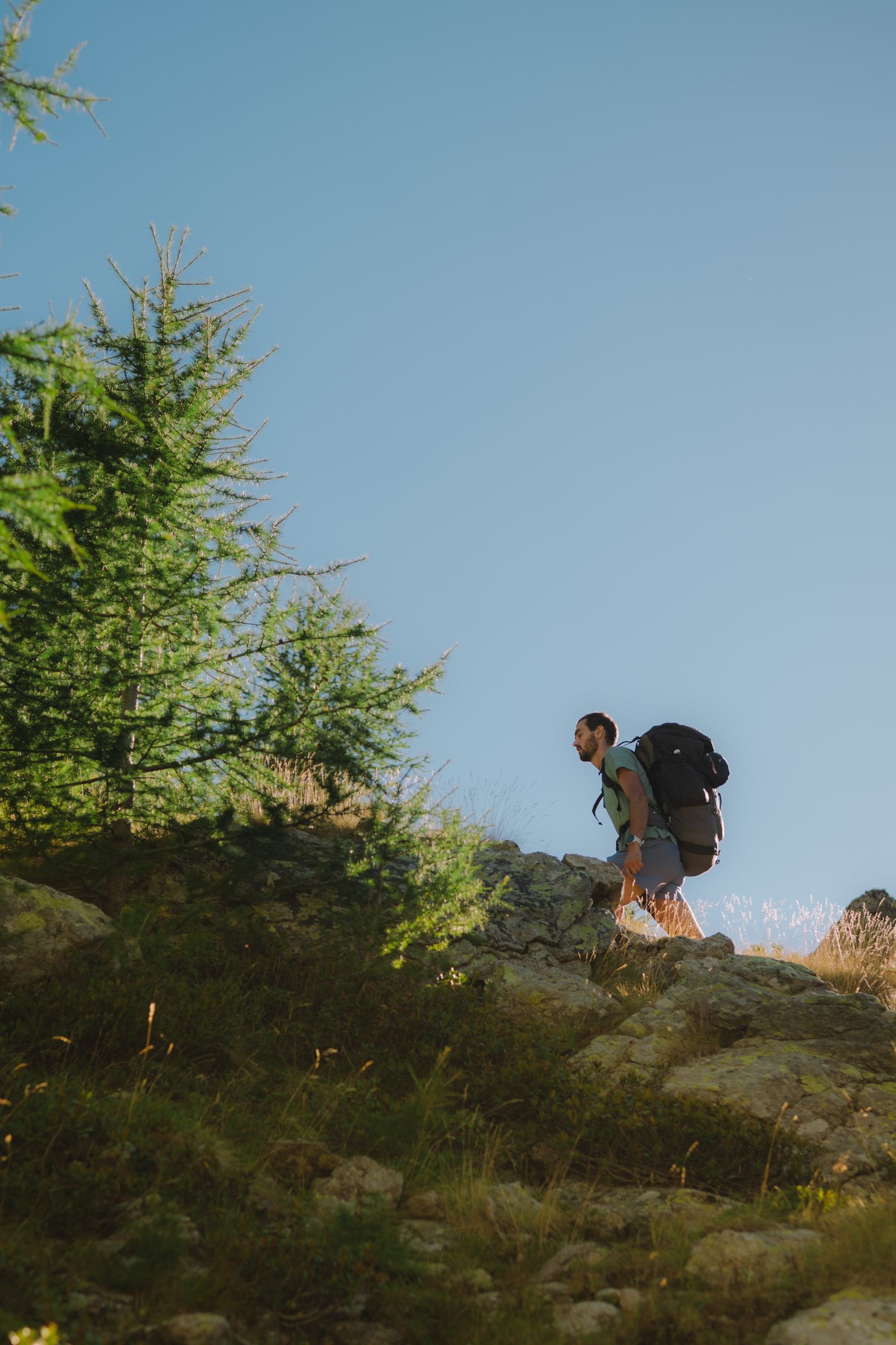 Un homme en randonnée escaladant une colline avec un grand sac à dos, entouré d'arbres sous un ciel bleu.