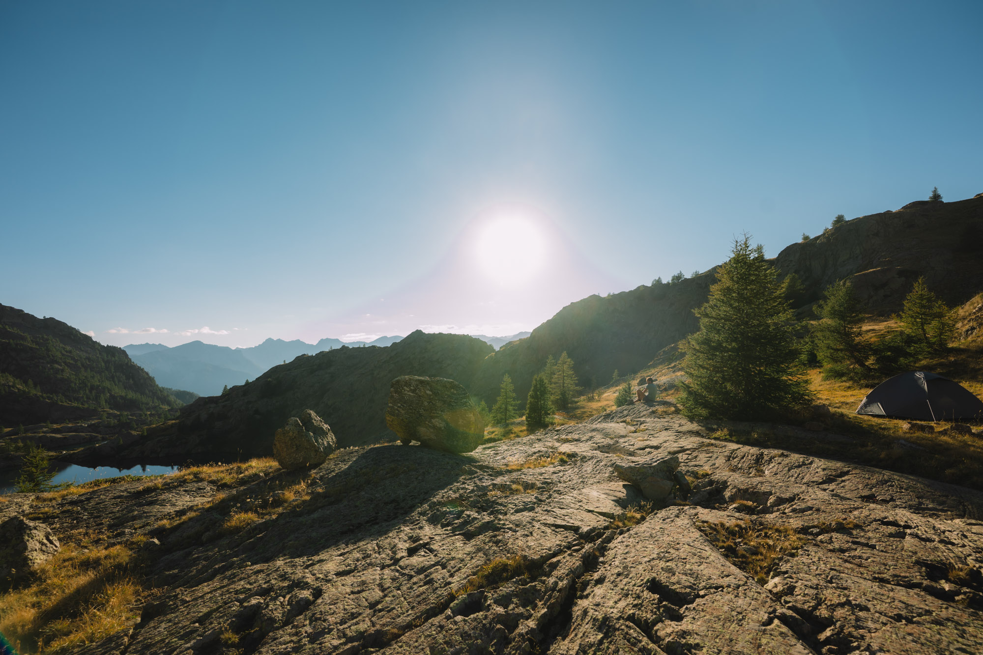 Paysage montagneux avec un lever de soleil, des rochers et des arbres, et une tente au premier plan.