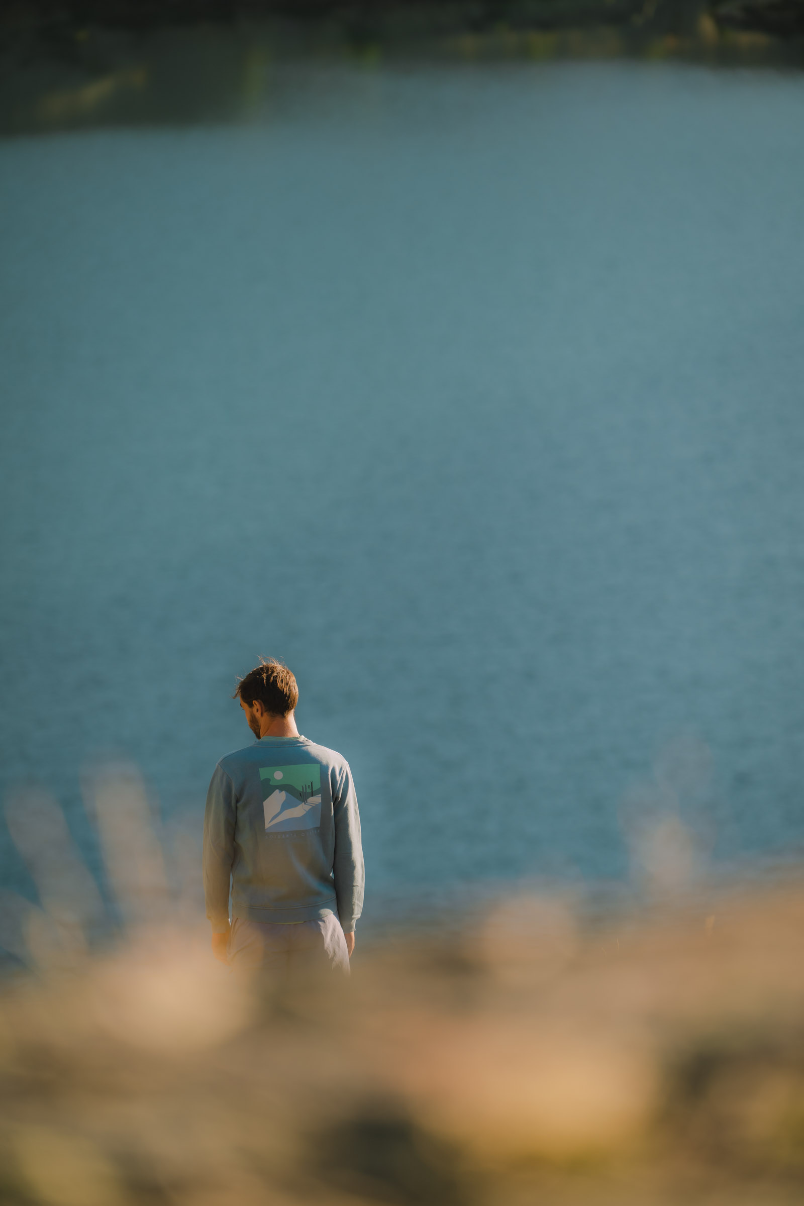 Un homme de dos, portant un sweat-shirt avec un motif de paysage, se tient au bord d'un lac tranquille.