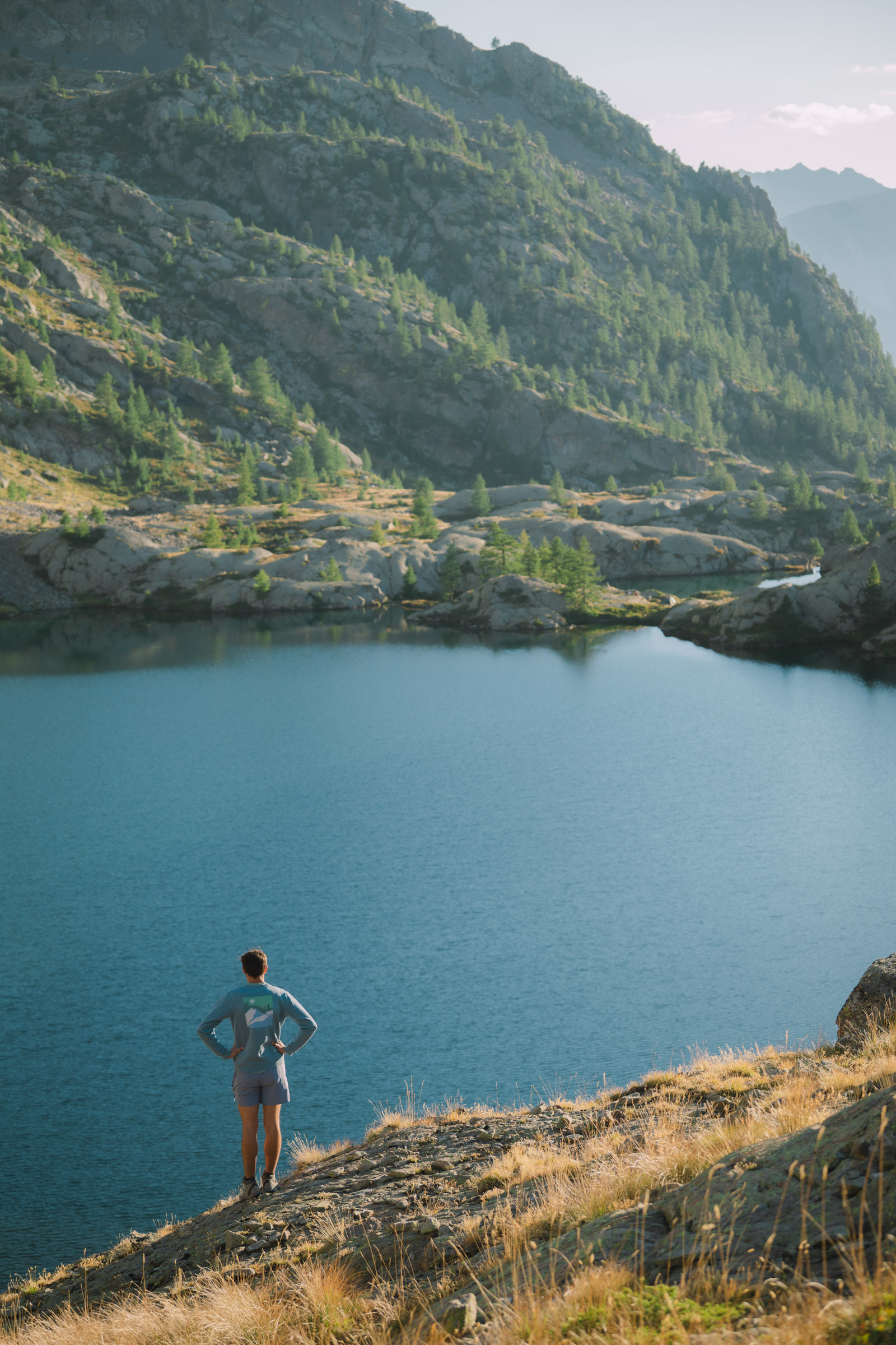Un randonneur debout au bord d'un lac, admirant un paysage montagneux avec des conifères et des rochers.