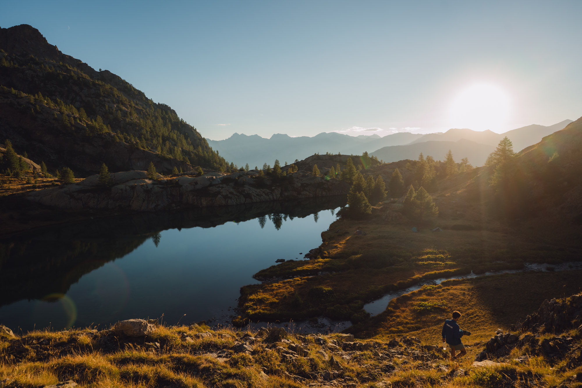 Un randonneur descendant une pente près d'un lac paisible, entouré de montagnes et de sapins, au coucher du soleil.